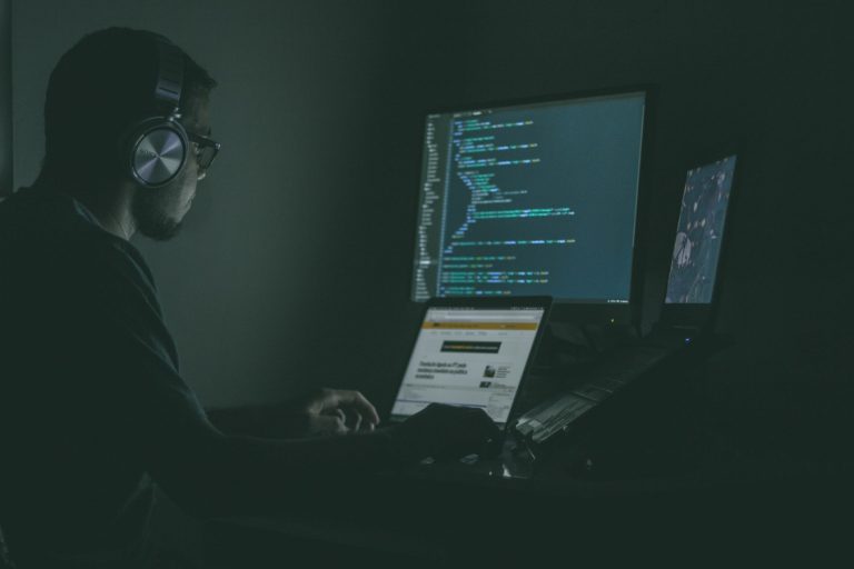 man listening to music while working on three computer screens writing malicious code.